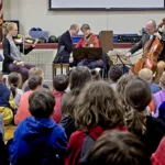 A string quartet performs on a stage in front of an audience of children. The musicians play violin, viola, and cello, while the children sit on the floor, watching attentively in a school gymnasium setting.