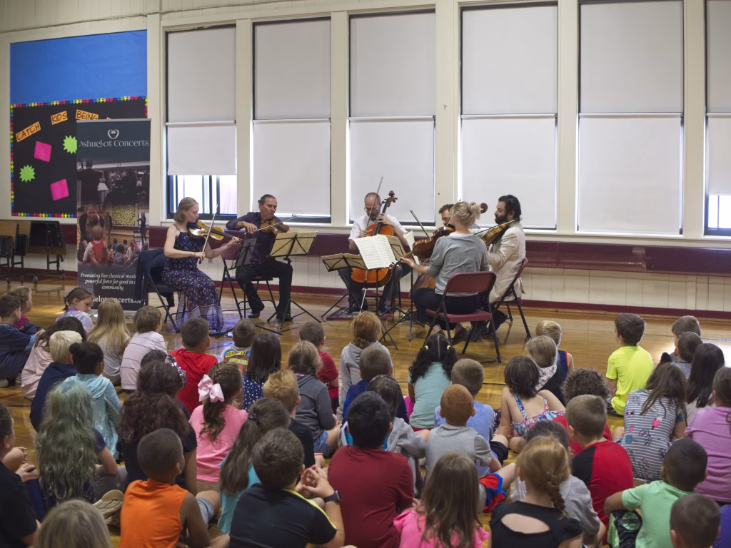 A string quintet performs for a group of young children seated on a gymnasium floor, with a banner and large windows in the background. The children watch the musicians attentively.