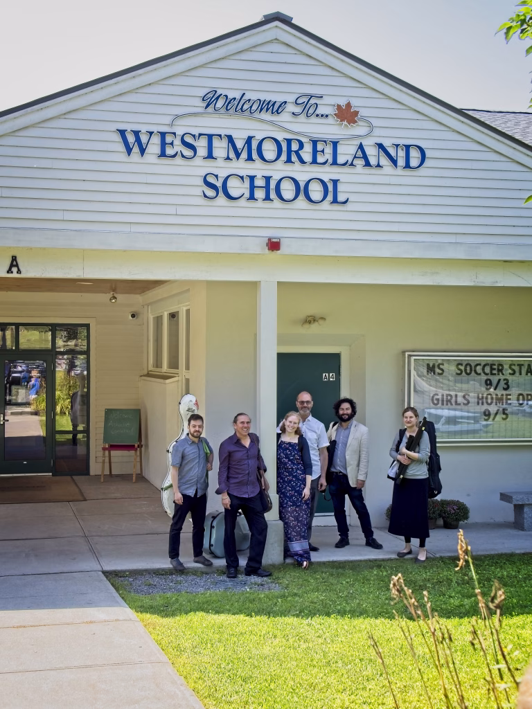 Six adults stand and pose in front of the entrance to Westmoreland School, which has a white exterior and a sign that reads “Welcome To... WESTMORELAND SCHOOL” above the door. A notice board is visible on the right.