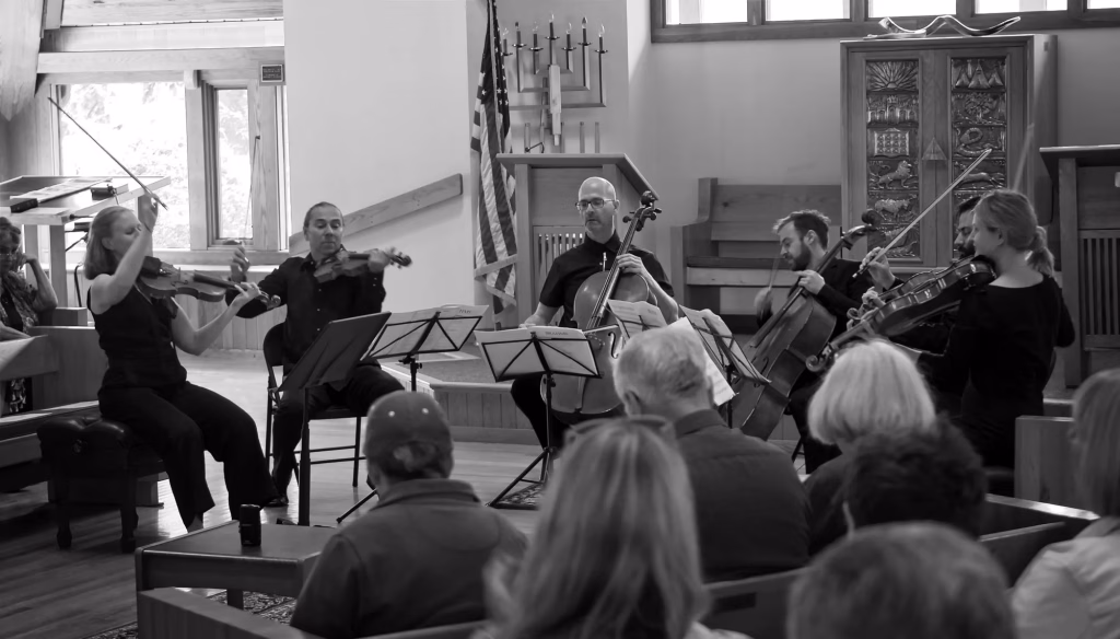 A black-and-white photo shows a string quintet performing indoors for an audience. The musicians play violins, viola, and cello while seated in front of music stands. The audience watches attentively.