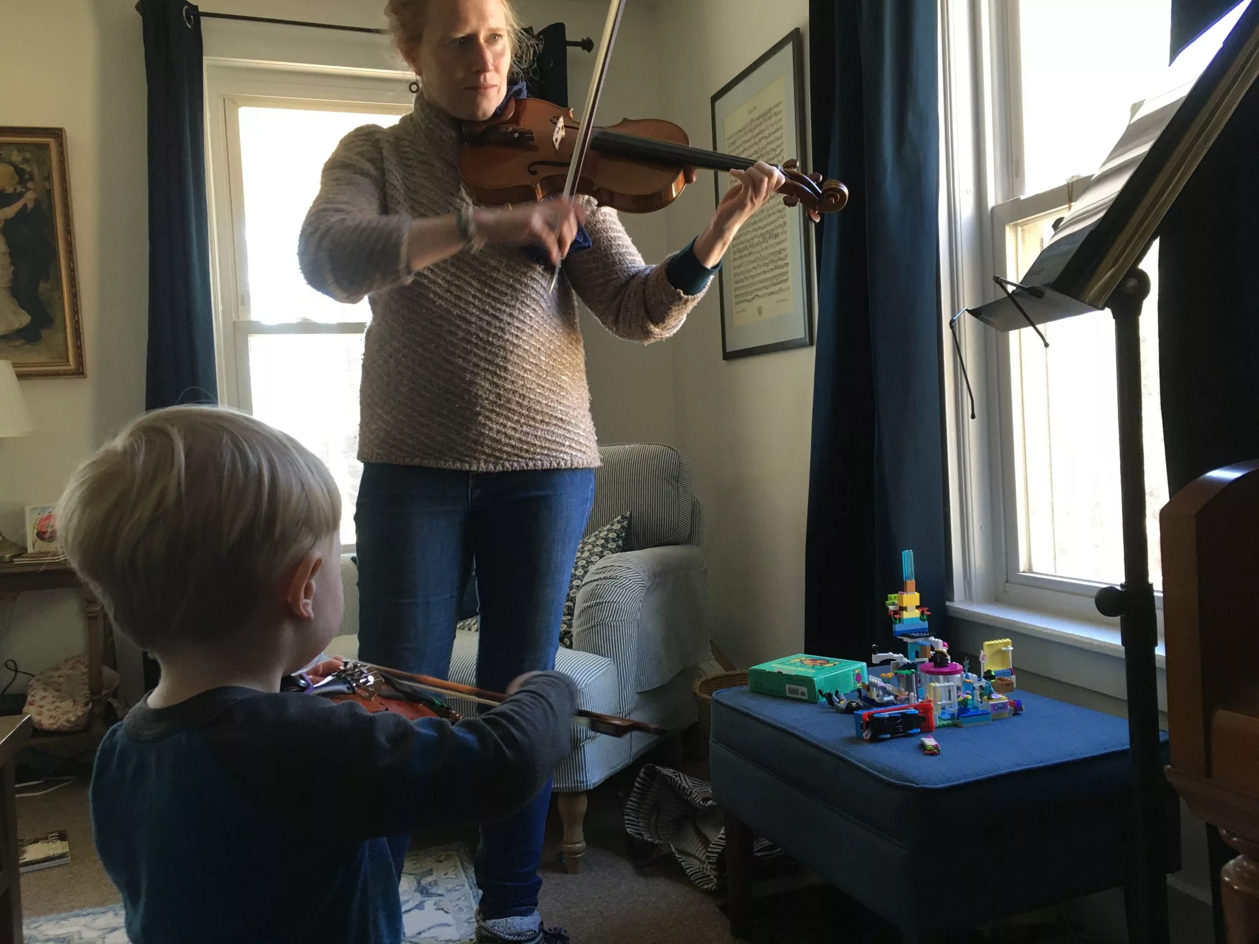 An adult and a child play violins together in a cozy living room. The adult follows sheet music on a stand, while the child mimics the posture. Sunlight streams through a window, and colorful toys are on a nearby ottoman.