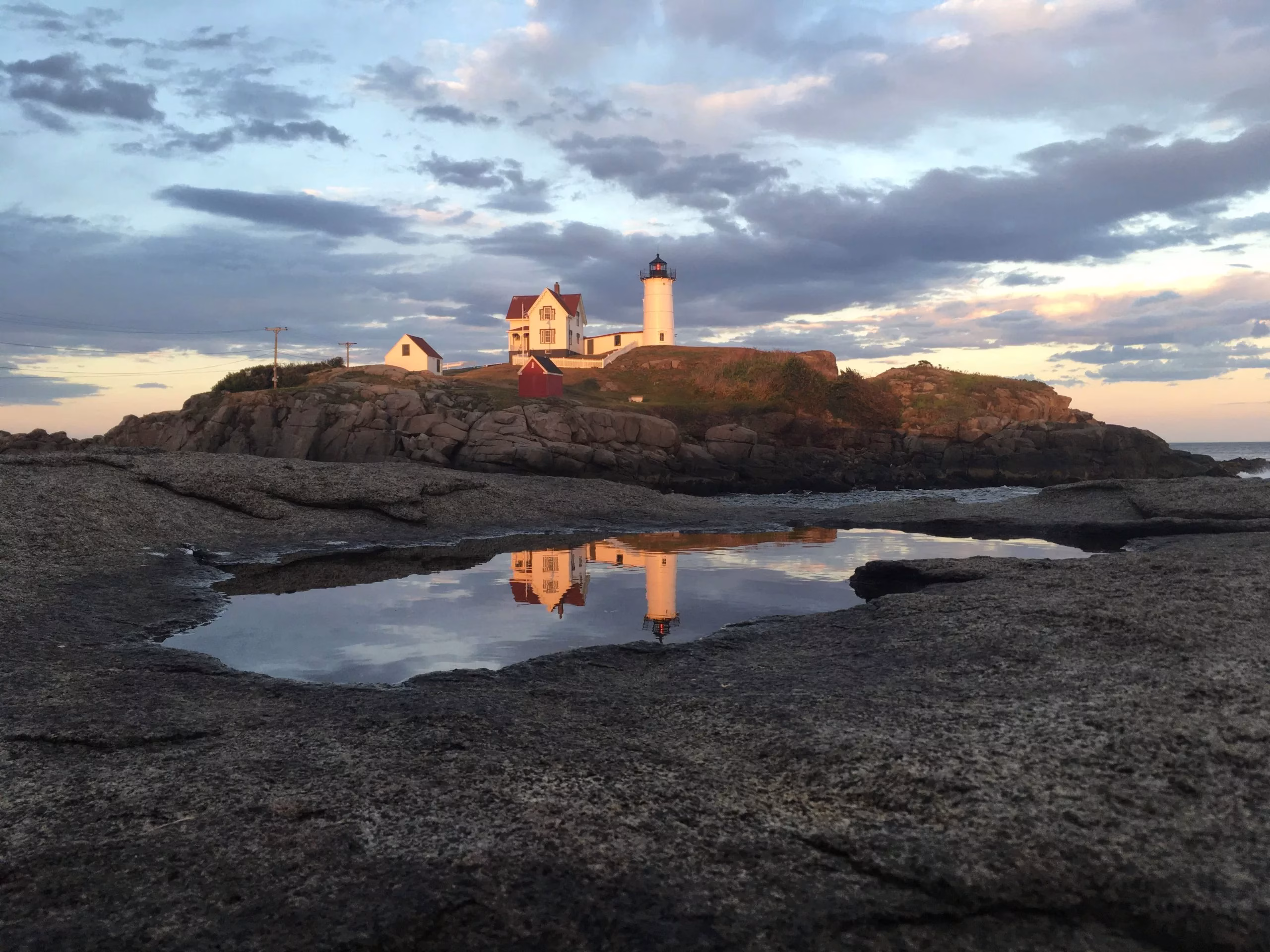 A lighthouse and house sit atop a rocky island, reflected in a tide pool below. The sky is partly cloudy with warm sunlight, and the ocean is visible to the right.