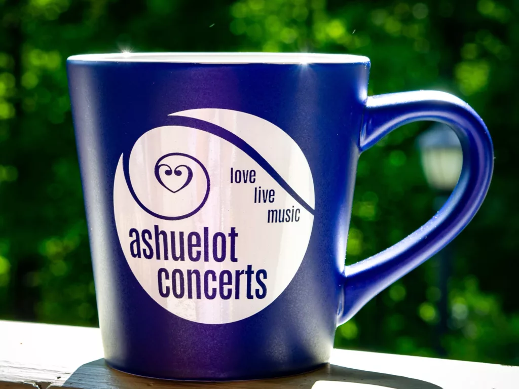 The Ashuelot Concerts Mug - Blue features a white logo and the words "ashuelot concerts" and "love live music," shown on a ledge with a blurred green outdoor background.