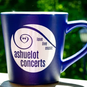 The Ashuelot Concerts Mug - Blue features a white logo and the words "ashuelot concerts" and "love live music," shown on a ledge with a blurred green outdoor background.