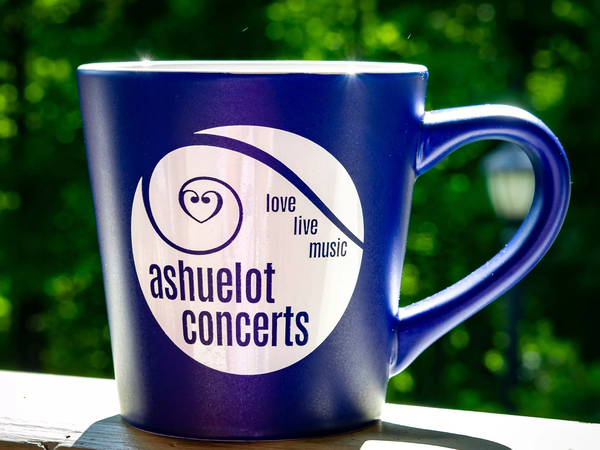 The Ashuelot Concerts Mug - Blue features a white logo and the words "ashuelot concerts" and "love live music," shown on a ledge with a blurred green outdoor background.