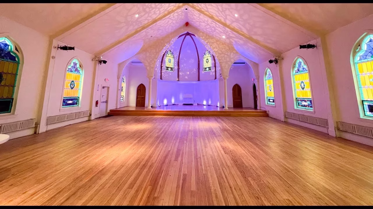 Spacious hall with wooden floors, stained glass arched windows, and a vaulted ceiling. A raised stage with purple lighting is at the far end, creating a serene and elegant atmosphere.