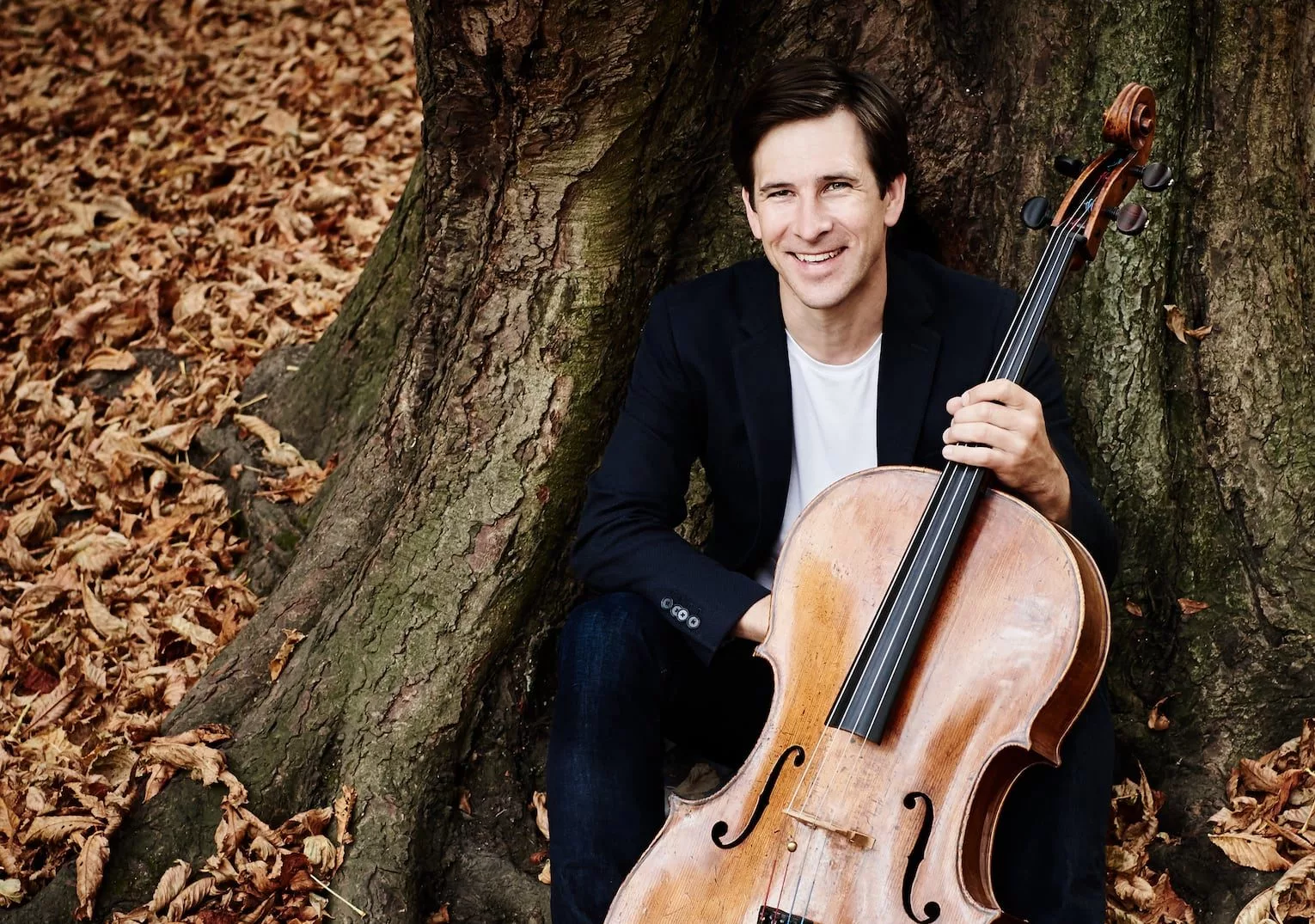 A man sits against a large tree trunk on fallen autumn leaves, smiling and holding a cello upright beside him. He wears a dark jacket and jeans.