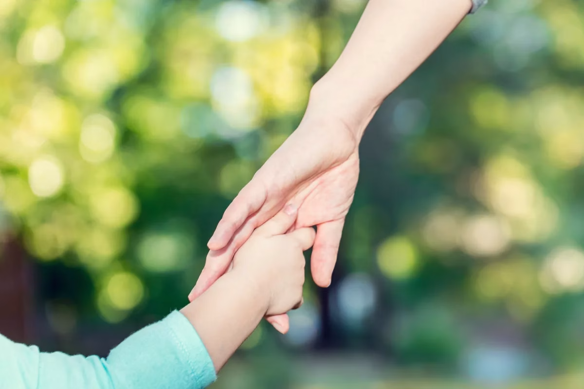 An adult hand gently holding a childs hand outdoors, with a blurred green background.