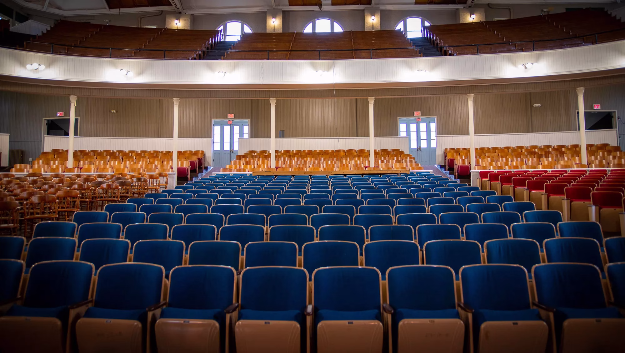 Rows of empty blue and brown seats face a stage in a spacious, well-lit auditorium with a balcony and large arched windows near the ceiling.