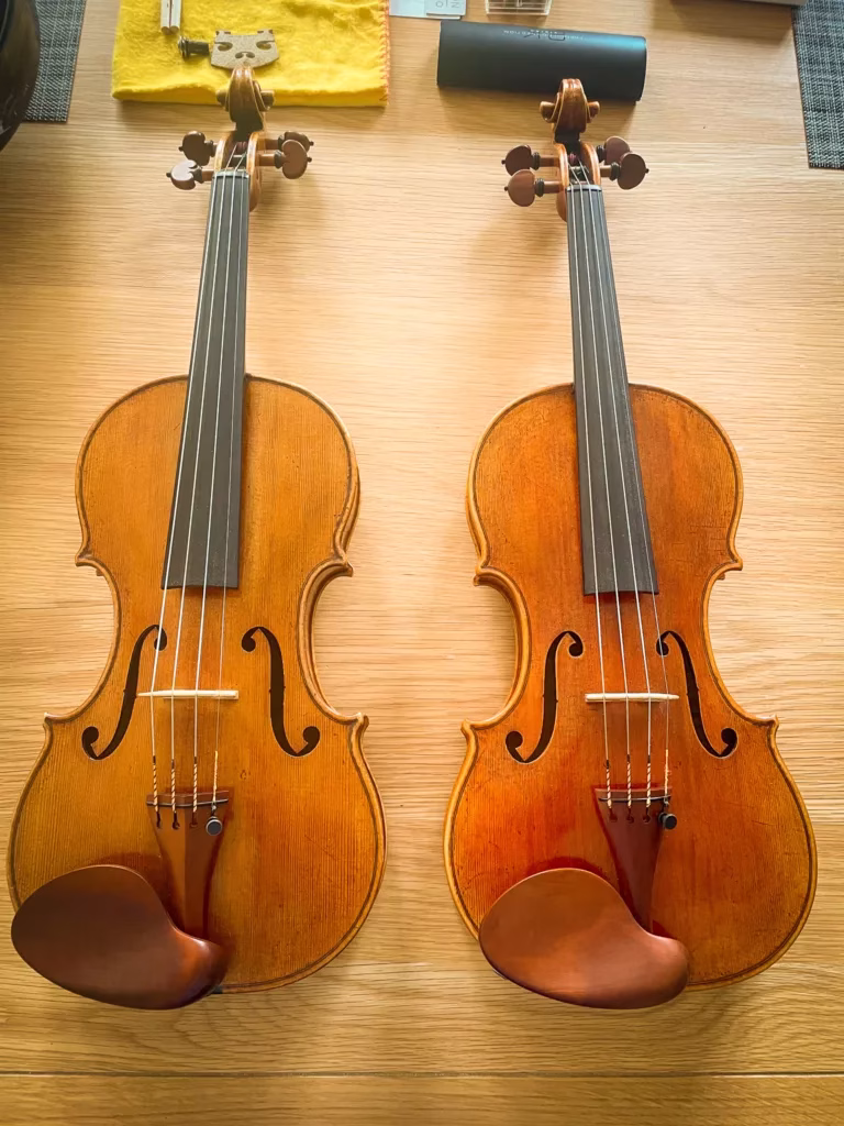 Two violins with brown wood finishes and dark fingerboards placed side by side on a light wooden surface, each with a chin rest and f-holes visible.