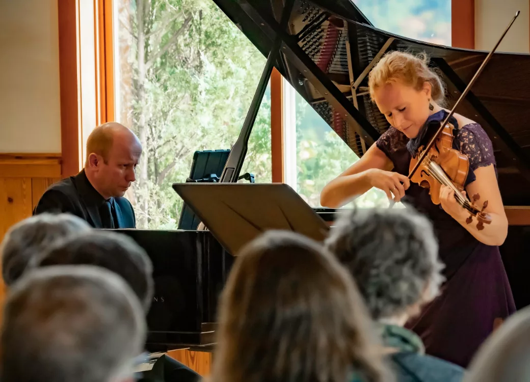 A woman plays the violin and a man plays the piano during a live indoor performance, with an audience watching and a grand piano in the background near large windows.