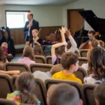 A group of children sit in chairs facing musicians with a violin, cello, and piano. Some children have their hands raised, and an adult stands at the front, engaging with the audience in a well-lit room.