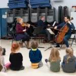 Three musicians—a violinist, a pianist, and a cellist—perform for a group of young children seated on the floor in a classroom or multipurpose room. Stacked chairs and mats are visible in the background.
