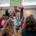 A group of children sit on the floor watching a woman play violin and a man play piano on stage in a school gymnasium. A “Destination Imagination” poster is displayed above the performers.