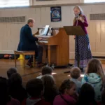 A man plays piano while a woman plays violin in front of an audience of children seated on the floor in a sunlit room with posters on the walls.