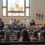 A small string quartet performs for an audience of children in a church, with a large stained glass window and altar visible behind them. An adult speaks while the musicians play and the children watch attentively.