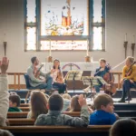 Four musicians perform with string instruments for children seated in church pews. A child in the audience raises their hand. A large stained glass window and candles are visible behind the musicians.