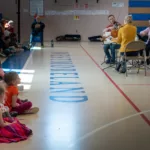 A string quartet performs in a gymnasium for a group of young children seated on the floor, with sunlight streaming in and WESTMORELAND visible on the gym floor.