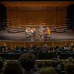 A small string quartet performs on a stage in an auditorium, with an audience seated and watching. The stage has risers in the background and flags on either side. The lighting focuses on the musicians.