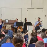 A string quartet and a speaker address a seated group of children in a gymnasium, with three musicians holding instruments and one person standing with a microphone.