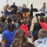 A group of musicians perform with string instruments and a piano in front of an audience of children seated on the floor, watching attentively.