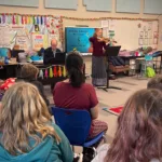 A woman plays the violin while a man plays piano in a colorful classroom, as children sit on blue chairs and watch. The classroom is decorated with posters, student artwork, and a U.S. flag.