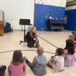 A woman kneels on the floor with a violin, smiling and talking to a group of young children seated around her in a school gymnasium. Musical instruments are in the background.