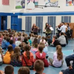 A string quartet performs for a large group of young children seated on the floor of a school gymnasium, while a few adults sit nearby. The gym features blue walls, sports equipment, and colorful artwork.
