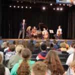 A quartet of musicians perform onstage with string instruments and a piano for an audience of students in a school gym. A man stands at a microphone, speaking to the crowd.