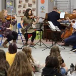 Four musicians perform for a group of seated children in a school gym. The musicians play string instruments and piano, while colorful artwork and posters decorate the wall behind them.
