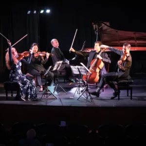 Five musicians perform on stage with string instruments and a grand piano behind them. They are seated in a semicircle, reading sheet music, illuminated by stage lights against a dark background.