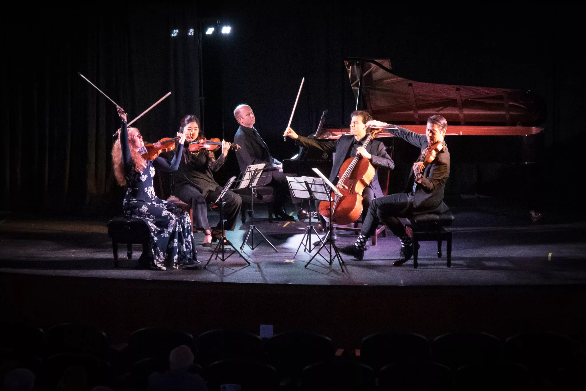 Five musicians perform on stage with string instruments and a grand piano behind them. They are seated in a semicircle, reading sheet music, illuminated by stage lights against a dark background.