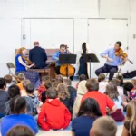 A string quartet and pianist perform for a large group of children seated on the floor in a gymnasium. The musicians face the audience, which consists of attentive, casually dressed kids.