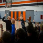 A woman stands in a gymnasium with her hands raised while a group of children, also raising their hands, face her. The gym features basketball hoops, orange and black accents, and equipment in the background.