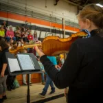 A person plays the violin in a gymnasium, reading sheet music on a stand. In the background, children and adults sit on bleachers, watching or talking to each other.