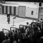 A man and a woman stand speaking in a gymnasium, addressing a seated audience of students. The gym has bleachers, a music stand, and TIGERS lettering on the floor. The scene is in black and white.