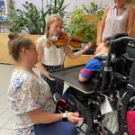 A young woman plays violin for a child in a wheelchair, while two women—one crouching and one standing—smile and watch in a bright indoor space with green plants in the background.