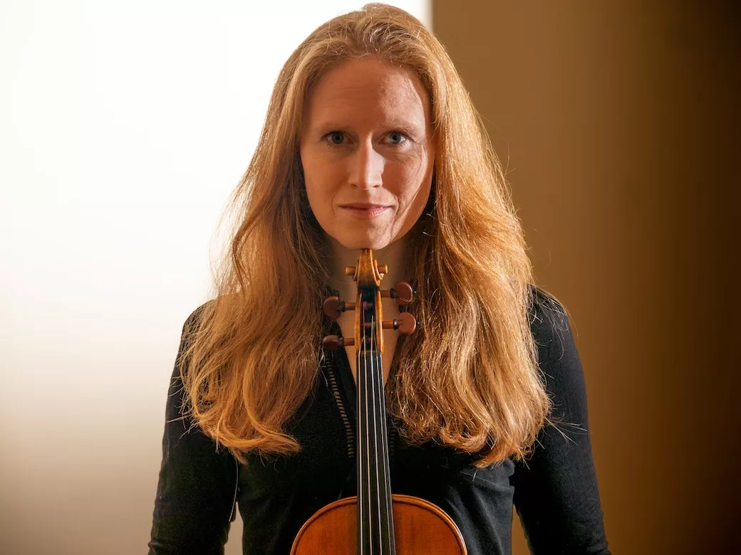 A woman with long red hair, wearing a black top, stands facing the camera while holding a violin upright in front of her. The background is softly lit with white and brown tones.