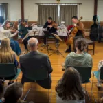 A string quartet performs for an audience seated in chairs in a sunlit room with wood floors, large windows, and a grand piano in the background. The listeners appear engaged, watching the musicians closely.