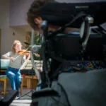 A woman plays the violin on a chair indoors, while a person in a wheelchair watches in the foreground. The setting appears to be a bright, community or healthcare space.