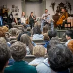 A group of musicians with string instruments perform and speak to an audience of students seated in rows, while a man stands to the side holding a microphone.