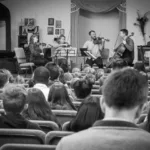 A string quartet performs onstage for an audience seated in rows inside a church or hall. The audience listens attentively, and musical stands and instruments are visible. The photo is in black and white.