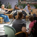 A string quartet performs for an audience in a brightly lit indoor space, with three violinists and a cellist. Two audience members in the foreground listen, one in a wheelchair. Green plants are in the background.