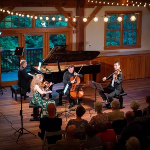 A string quartet and pianist perform on a wooden stage under string lights, with an audience seated in front. The venue has large windows and warm wooden paneling.