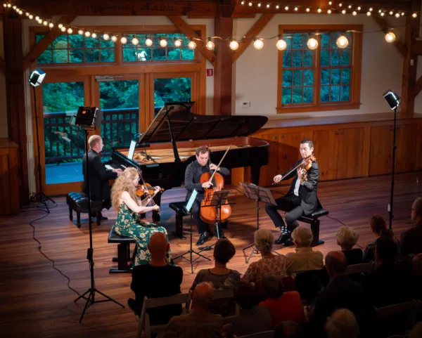 A string quartet and pianist perform on a wooden stage under string lights, with an audience seated in front. The venue has large windows and warm wooden paneling.