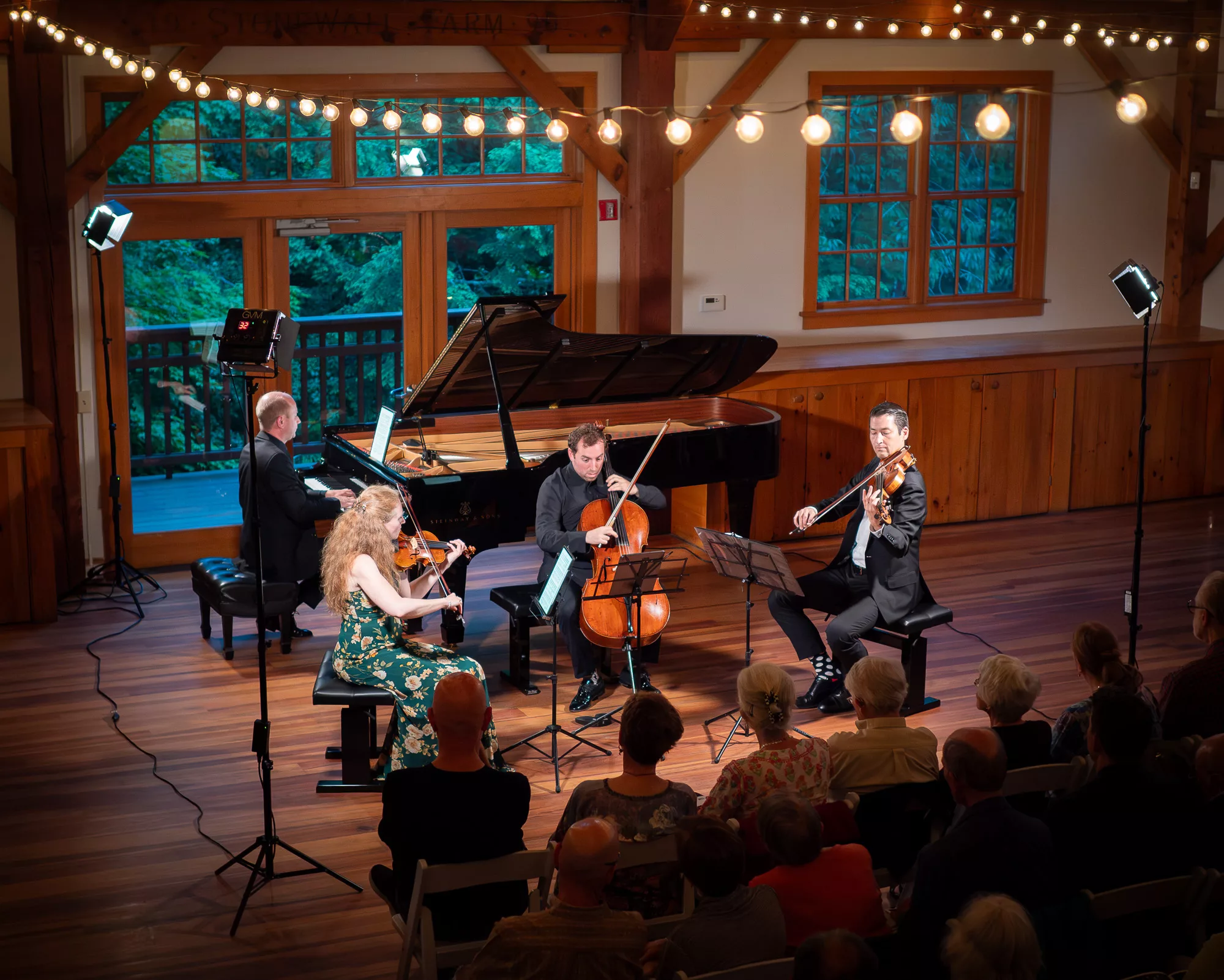 A string quartet and pianist perform on a wooden stage under string lights, with an audience seated in front. The venue has large windows and warm wooden paneling.