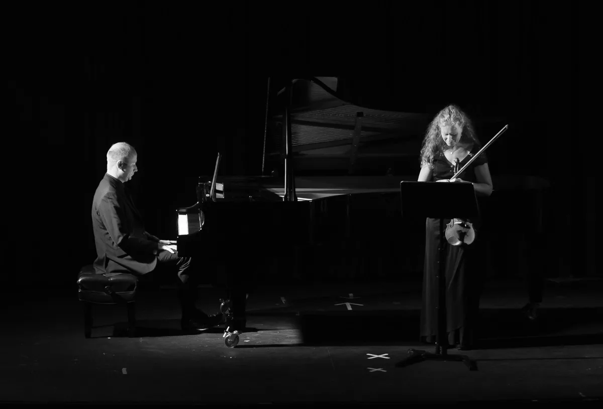 A man plays a grand piano while a woman stands nearby playing a violin. Both musicians are illuminated on a dark stage, with sheet music on a stand in front of the violinist.