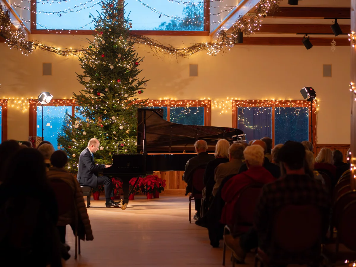 A man in a suit plays a grand piano on stage before an audience in a hall decorated with string lights and a large Christmas tree, creating a festive atmosphere.