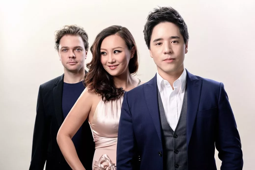 Three people posing together against a light background. A man in a blue suit and white shirt stands in front, a woman in a pale pink dress smiles in the middle, and another man in a black jacket stands behind them.