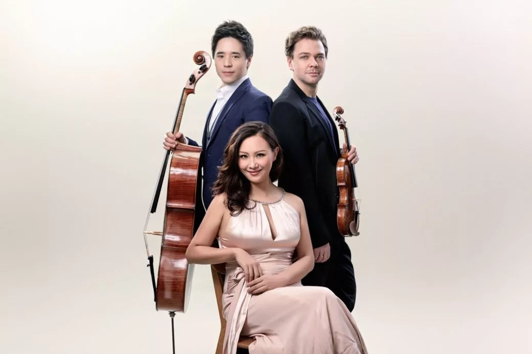 Three musicians pose against a plain background: a woman in a pale pink dress sitting in front, with two men standing behind her, one holding a cello and the other holding a violin, all dressed formally.